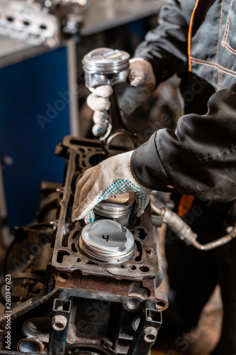 Close-up Car mechanic holding a new piston for the engine, overhaul.. Engine on a repair stand with piston and connecting rod of automotive technology. Interior of a car repair shop.