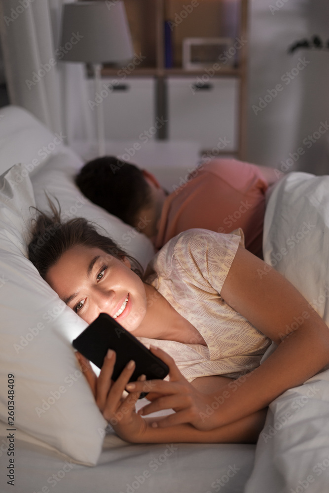 technology, internet addiction and cheat concept - happy smiling woman using smartphone at night while her boyfriend is sleeping technology, internet addiction and cheat concept - happy smiling woman using smartphone at night while her boyfriend is sleeping