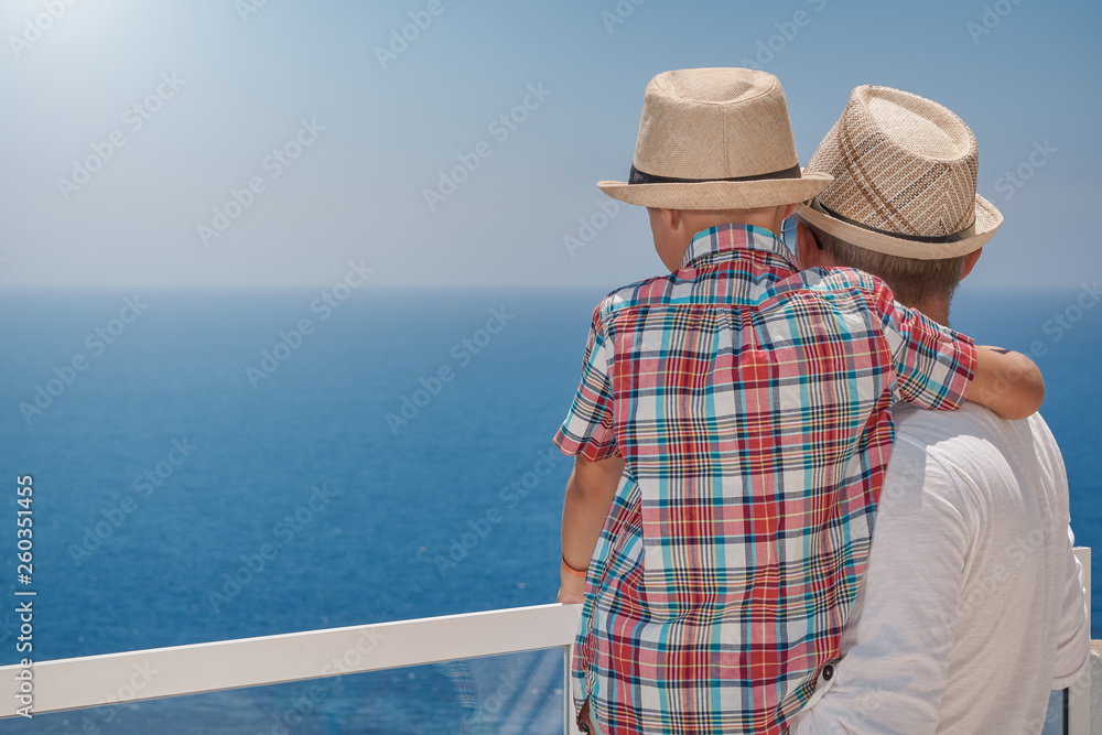 Son and dad relaxing on hotel balcony against sea. Father is holding son on hands.