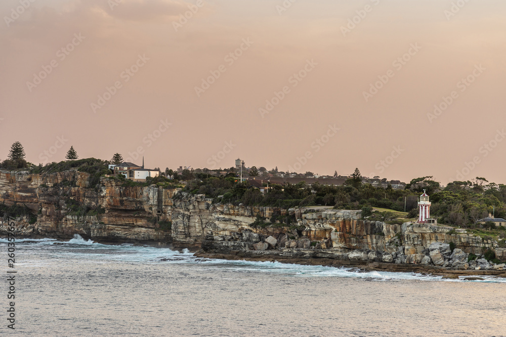 Sydney, Australia - February 12, 2019: South Head cliffs at gate between Tasman Sea and Sydney Bay during sunset. Cloudless orange sky. Gray water. Crashing waves. Warm brown rocks Hornby lighthouse.