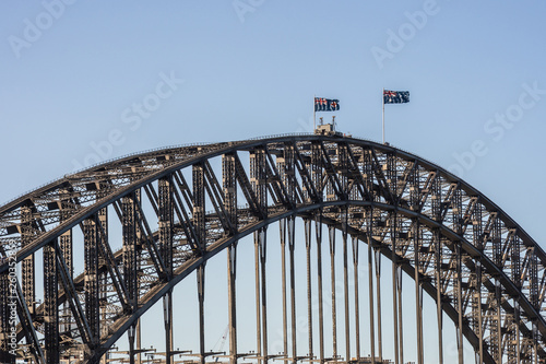 Sydney, Australia - February 12, 2019: Closeup of Harbour bridge span during sunset. Black metal, Flags and light blue sky. © Klodien