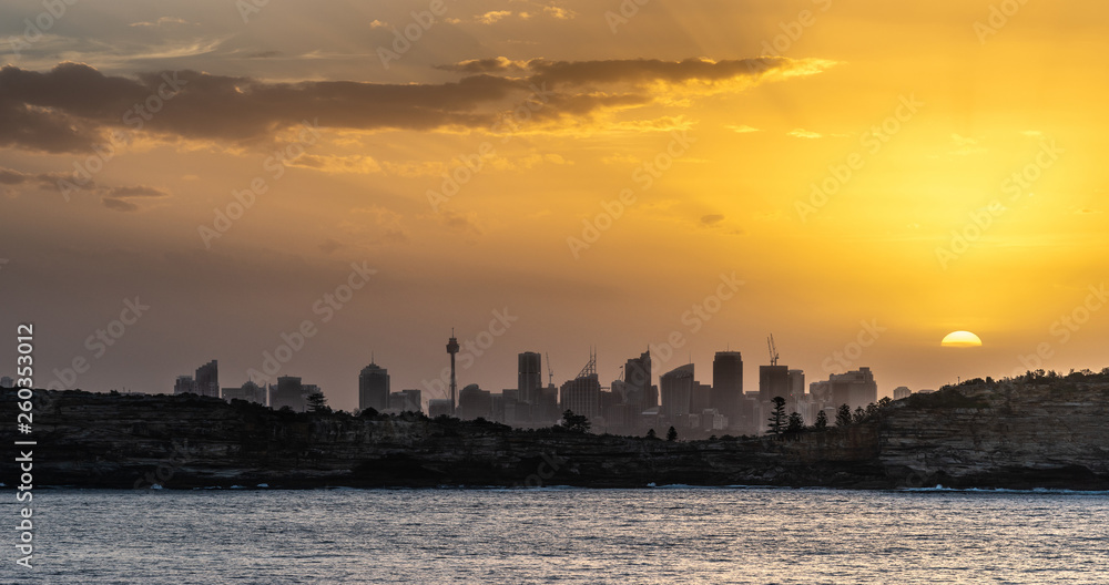 Sydney, Australia - February 12, 2019: Sunset over city skyline seen from Tasman Sea. Shoreline rocky cliffs. Yellow brown sky, sun rays. 5 of 5.