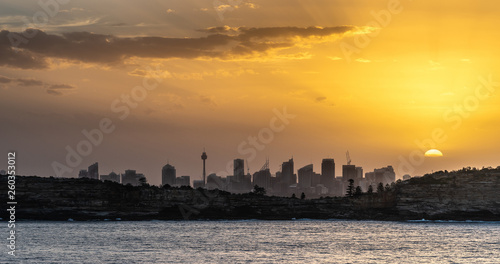 Sydney  Australia - February 12  2019  Sunset over city skyline seen from Tasman Sea. Shoreline rocky cliffs. Yellow brown sky  sun rays. 5 of 5.