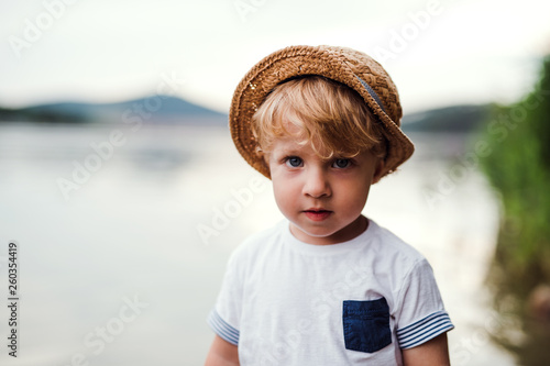 A small toddler boy standing outdoors by a river in summer, looking at camera. photo