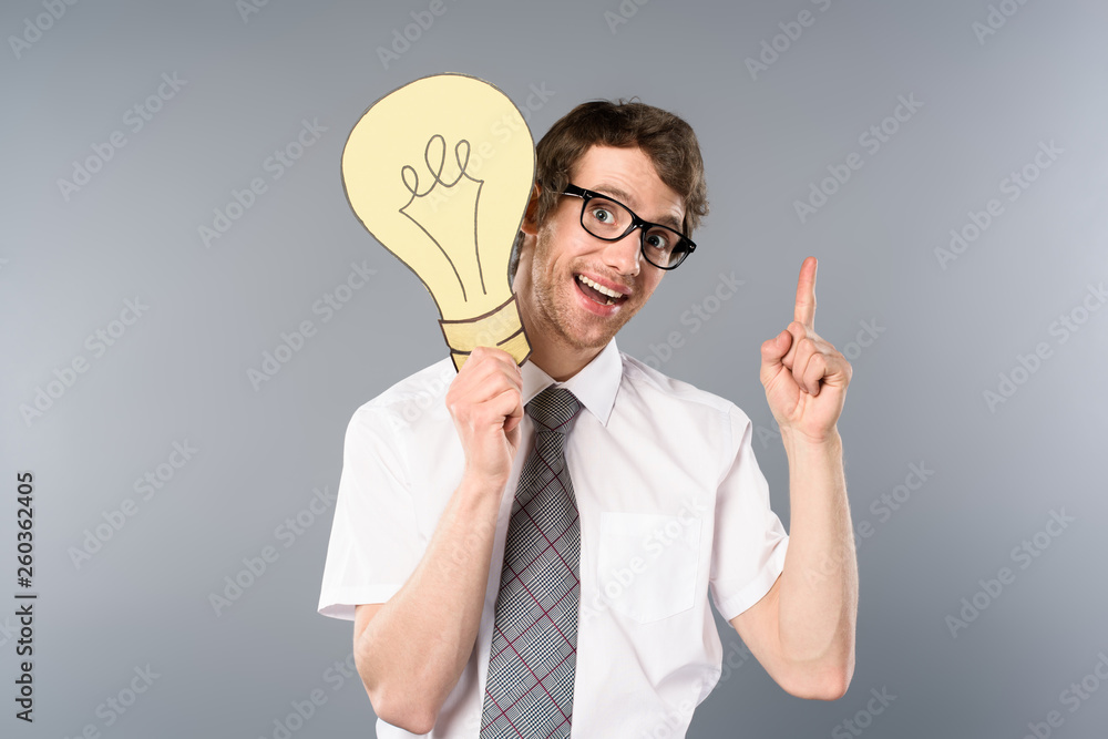 smiling businessman in glasses holding yellow paper cut light bulb and showing idea gesture on grey background smiling businessman in glasses holding yellow paper cut light bulb and showing idea gesture on grey background