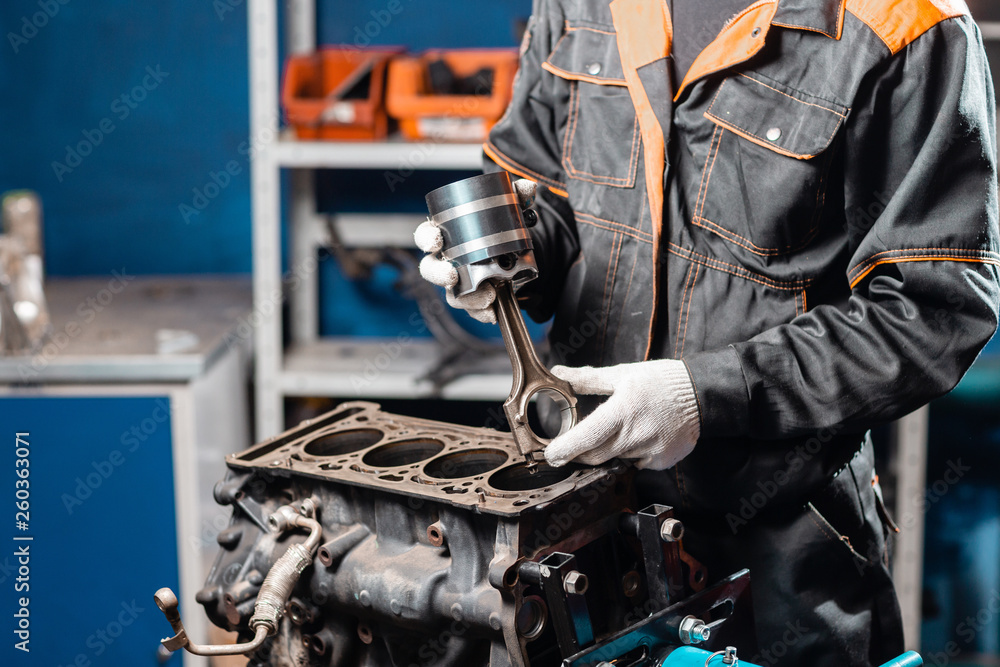Car mechanic holding a new piston for the engine, overhaul.. Engine on a repair stand with piston and connecting rod of automotive technology. Interior of a car repair shop. Stock Photo |