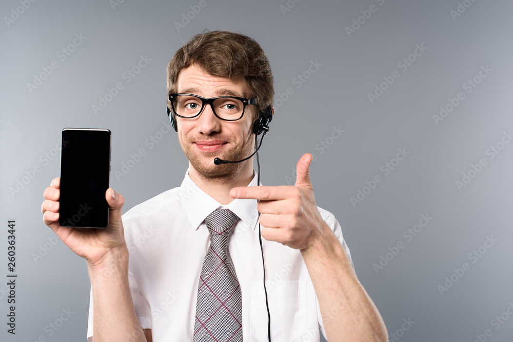 positive call center operator in glasses and headset pointing with finger at smartphone with blank screen positive call center operator in glasses and headset pointing with finger at smartphone with blank screen