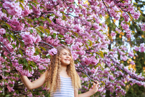 Adorable little girl with curly hair in pink dress walking in blossom cherry garden in sunny spring day