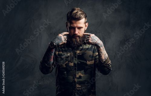 Portrait of a stylish bearded guy with tattooed hands in the military shirt. Studio photo against dark wall