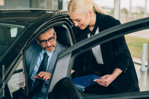 Car saleswoman and male customer in driver’s seat of new car in car dealership showroom