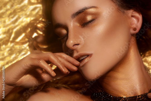 Gold Fashion Makeup, Art Beauty Face and Lips Make Up in Golden brocade. Fashion golden skin Woman face portrait. Gold jewellery, jewelry, accessories, earrings bracelet. Golden shadows. Girl lies.