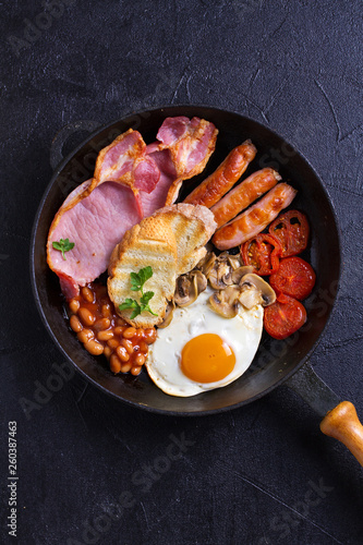 Full English or Irish breakfast with sausages, bacon, eggs, tomatoes, mushrooms and beans on black background