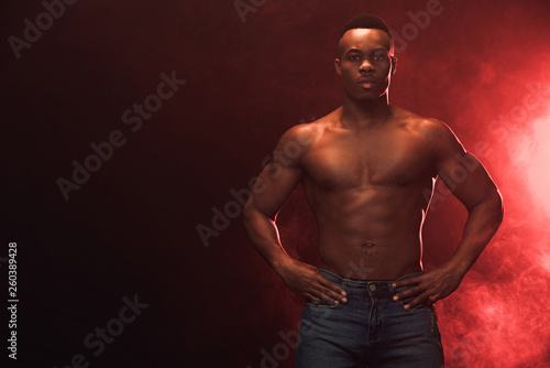 sexy muscular african american man in denim looking at camera on dark with red light and copy space