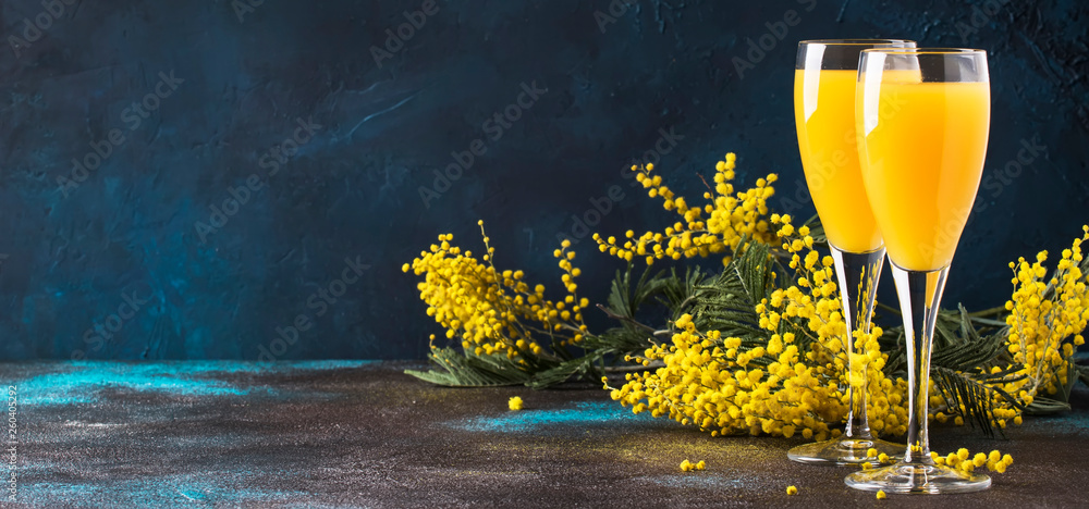 Classic alcohol cocktail mimosa with orange juice and cold dry champagne or sparkling wine in glasses, blue stone background with yellow flowers, copy space, spring mood, selective focus - obrazy, fototapety, plakaty 