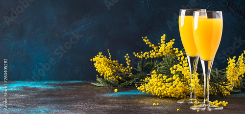 Classic alcohol cocktail mimosa with orange juice and cold dry champagne or sparkling wine in glasses, blue stone background with yellow flowers, copy space, spring mood, selective focus