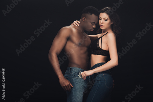 attractive woman embracing shirtless african american man and holding condom isolated on black