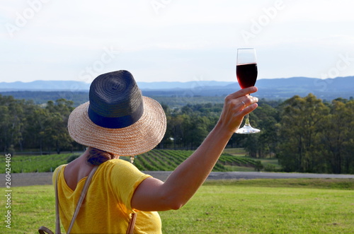 A woman wearing a hat and holding a glass with red sparkling wine admiring a beautiful landscape of winery fields, Hunter Valley, Australia