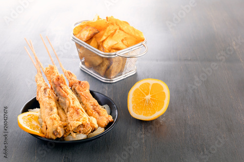 baked fish on skewers in a black plate, french fries and lemon on a wooden table. Copy space