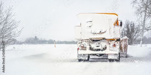 Orange plough truck on snow covered road, gray sky and trees in background, view from back - winter road maintenance