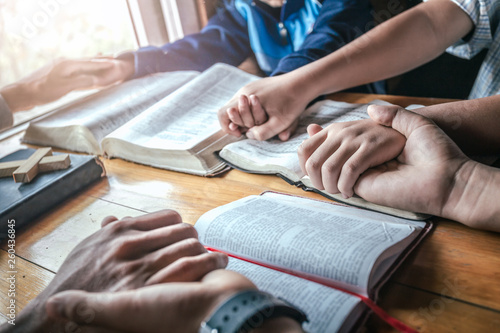 Christian group holding hands and praying together around wooden table with open bible page at home, prayer meeting concept.