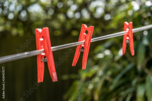 Three red plastic clothes pegs on a clothes line against the background of blurred green bush.