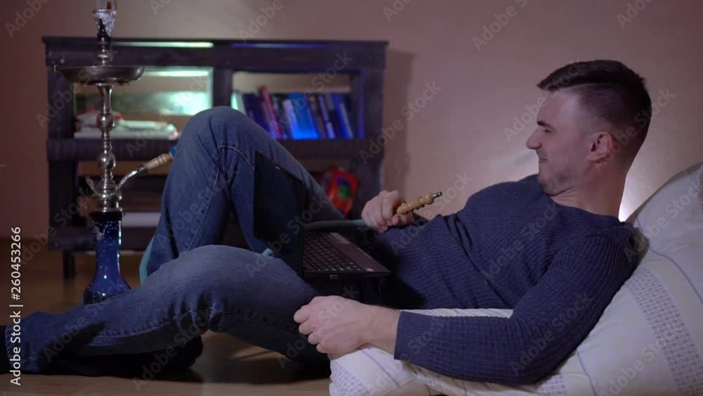 The young guy smokes a hookah, exhales smoke and watches the video on the laptop, he does not like. A young man in the smoke. Young guy on the pillow on the background of the bookcase. The young guy smokes a hookah, exhales smoke and watches the video on the laptop, he does not like. A young man in the smoke. Young guy on the pillow on the background of the bookcase.