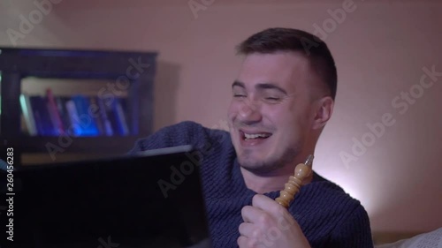 The young guy smokes a hookah, exhales smoke and watches over the laptop, the guy laughs and feels delight. On the background of a bookcase, close-up. photo