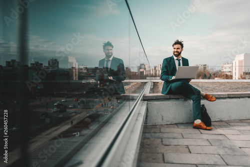 Young smiling cheerful Caucasian bearded businessman in formal wear using laptop while sitting on rooftop. At first they will ask you why you are doing it, later they will ask you how you did it.