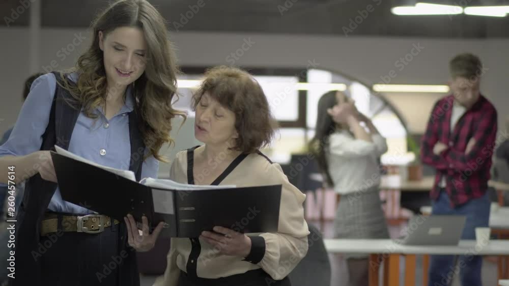 Two women discussing project standing in the office with big paper folder. Positive bearded man in casual clothes pass by on his bike drinking coffee. Active working life in office. Workplace Two women discussing project standing in the office with big paper folder. Positive bearded man in casual clothes pass by on his bike drinking coffee. Active working life in office. Workplace
