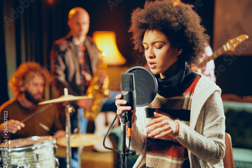 Mixed race woman singing. In background band playing instruments. Home studio interior.