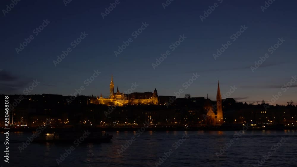 Wide shot of Budapest by night, Danube river bank with Matthias church on Buda, as a ship passing by Wide shot of Budapest by night, Danube river bank with Matthias church on Buda, as a ship passing by