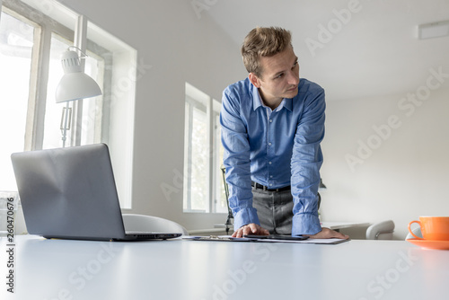 Handsome young businessman leaning on his office desk as he browses the digital tablet