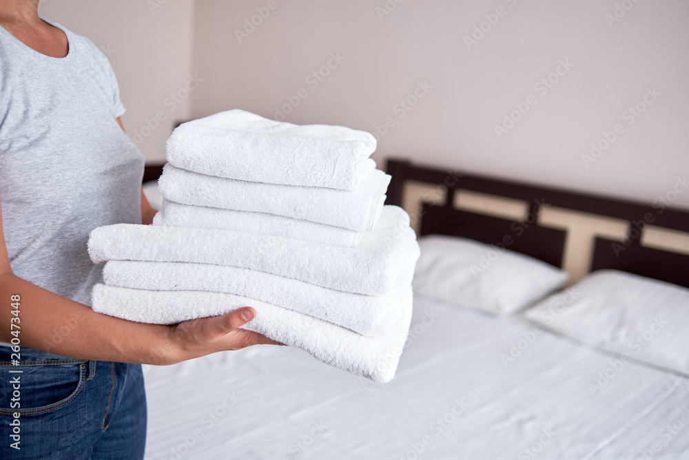 Woman holding stack of white clean bath towels in bedroom interior, copy space. Close up hands of hotel maid with towels. Room service Woman holding stack of white clean bath towels in bedroom interior, copy space. Close up hands of hotel maid with towels. Room service