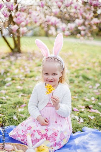 Little cute girl sitting on the grass near the magnolia. A girl dressed as an Easter bunny holds a flower and an egg.