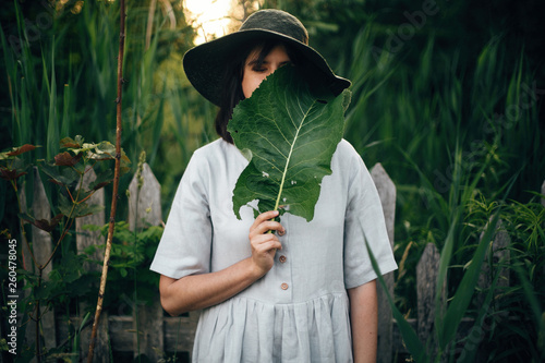 Stylish girl in linen dress holding big green leaf at face at wooden fence and grass. Portrait of boho woman in hat posing with leaf in summer countryside in evening. Atmospheric moment