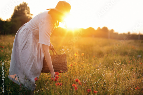 Stylish girl in linen dress gathering flowers in rustic straw basket, walking in poppy meadow in sunset. Boho woman in hat relaxing in warm evening sunlight in summer field photo