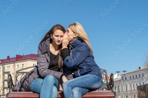 Portrait of two young attractive happy female friends women brown-haired and blonde overhear a whisper secret (mystery) on the street in a park on a bench. concept of gossip and privacy.