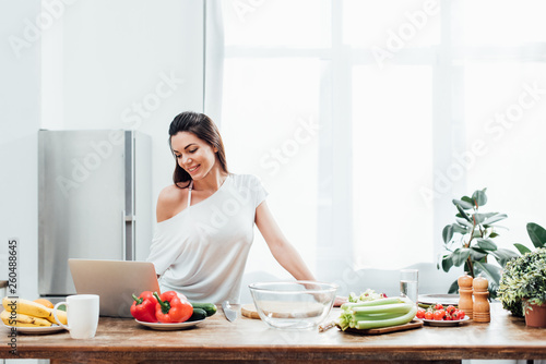 Attractive young woman using laptop at table in kitchen