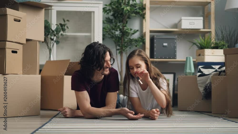 Cute girl is giving keys to handsome boyfriend and kissing him after moving to new house. People are lying on carpet together surrounded by carton boxes. Cute girl is giving keys to handsome boyfriend and kissing him after moving to new house. People are lying on carpet together surrounded by carton boxes.