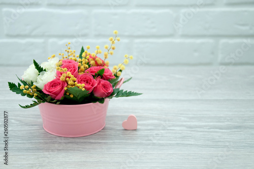 Fototapeta Naklejka Na Ścianę i Meble -  Creative bouquet of flowers with a pink heart against the background of a white loft, on a wooden table. Spring bright bouquet in a pink decorative bucket.