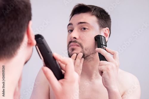 A young guy shaves in the bathroom. The guy shaves his beard with a trimer. Care beard.