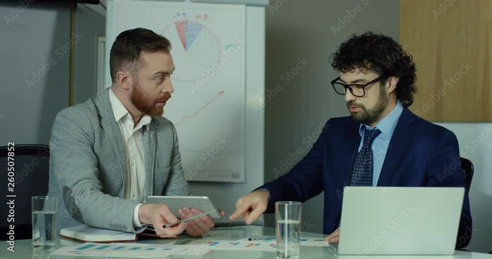 Two Caucasian attractive men xo-workers sitting together in the office and working on the laptop and tablet computer on their common project. Two Caucasian attractive men xo-workers sitting together in the office and working on the laptop and tablet computer on their common project.