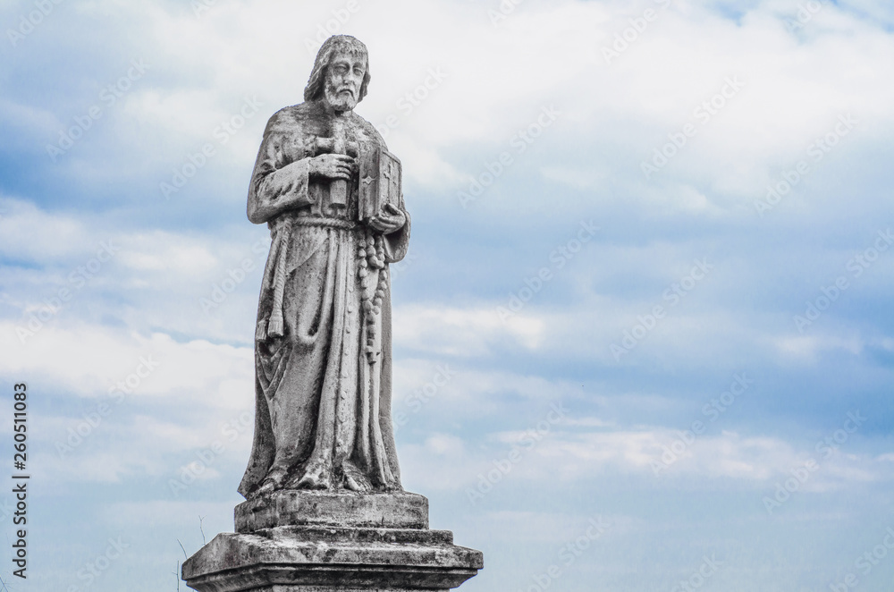 Ancient religious statue in the old cemetery on the background sky with clouds. Jesus with bible
