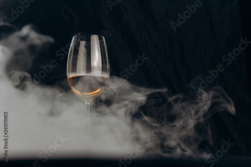 a glass of white wine on a black background. the smoke from the hookah envelops the glass. rest, holiday. alcoholic drink closeup. white fog in a glass. fog