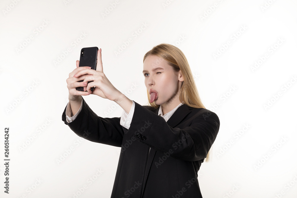 Young attractive female in dark suit takes photos using mobile phone, close one eye and showing tongue Young attractive female in dark suit takes photos using mobile phone, close one eye and showing tongue