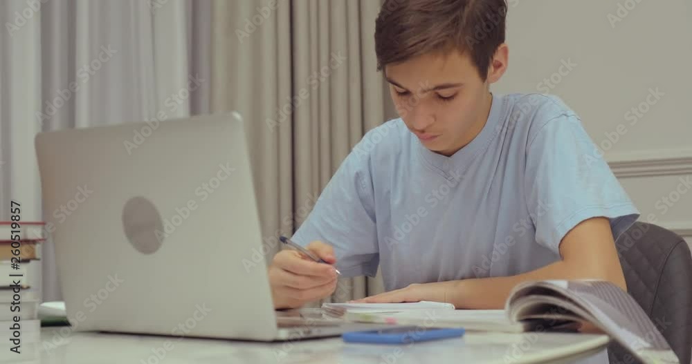 Teen boy doing school lessons at home. Student writes the homework. Child learns. Boy using laptop. Student writes with a pen in a notebook. Teen boy doing school lessons at home. Student writes the homework. Child learns. Boy using laptop. Student writes with a pen in a notebook.