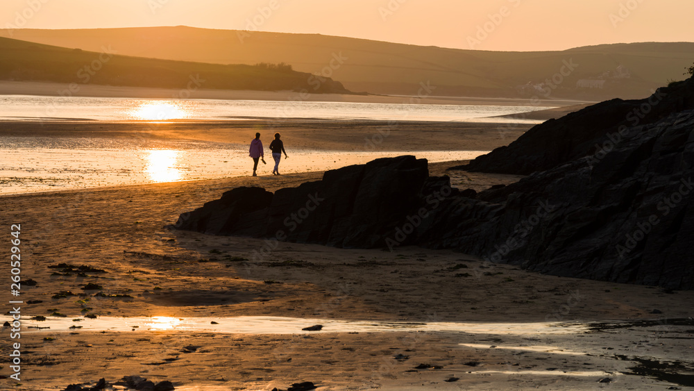 Rock, Cornwall, England UK: Two females in middle distance walking barefoot along the shoreline of the Camel estuary, in silhouette with late summer sun reflected in the water. Rock, Cornwall, England UK: Two females in middle distance walking barefoot along the shoreline of the Camel estuary, in silhouette with late summer sun reflected in the water.