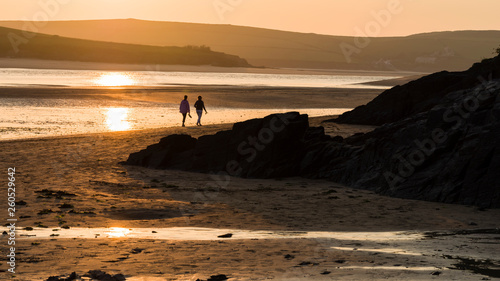 Rock, Cornwall, England UK: Two females in middle distance walking barefoot along the shoreline of the Camel estuary, in silhouette with late summer sun reflected in the water.