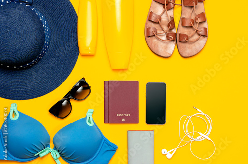 Female summer beach accessories on yellow background. Concept of travel vacation sea. Blue hat swimming suit leather sandals sunglasses sunscreen passport phone headphones Flat lay top view