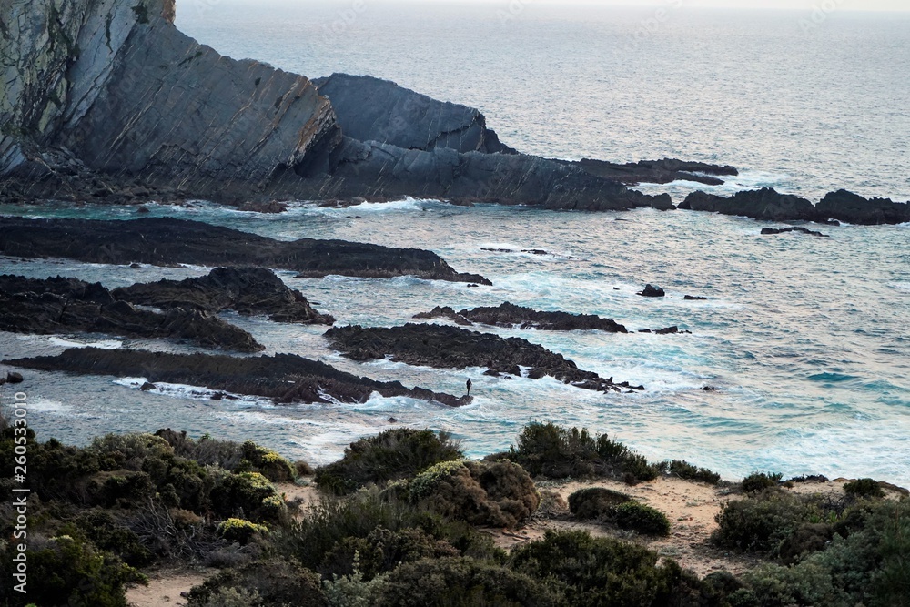 The man standing on the cliffs in the water, seen from far away, big waves. Rota Vicentina, Alentejo, Portugal